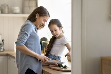 Little girl sit on kitchen countertop help mum chopping...