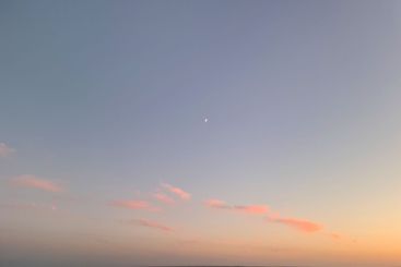 Little moon hangs in the sky over the ocean during dusk,...