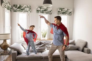 Cheerful dad and son in red superhero mantles dancing...