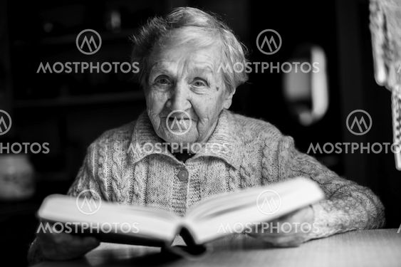 Elderly woman sitting at a table reading a book. Black-and-white contrast portrait.