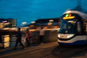 Tram passing by river and pedestrians at dusk in city...