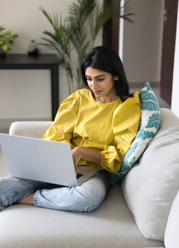 Indian woman using laptop seated on cozy sofa at home