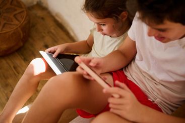 Siblings using smartphone and tablet in a cozy indoor...