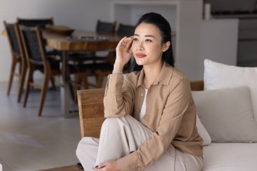 Positive dreamy young Asian woman sitting on sofa at home