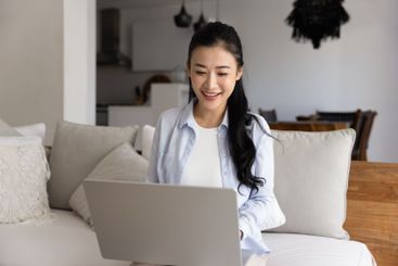 Happy Asian freelancer girl using laptop on home sofa