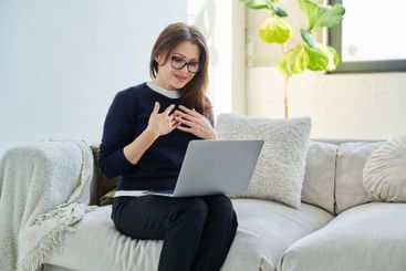 Middle aged woman sitting at home on sofa using laptop...