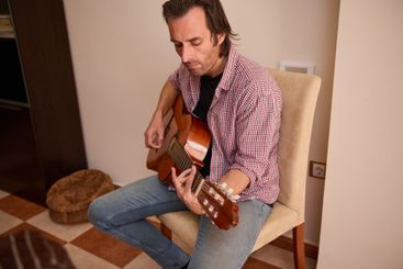 Man playing acoustic guitar while seated indoors on a chair