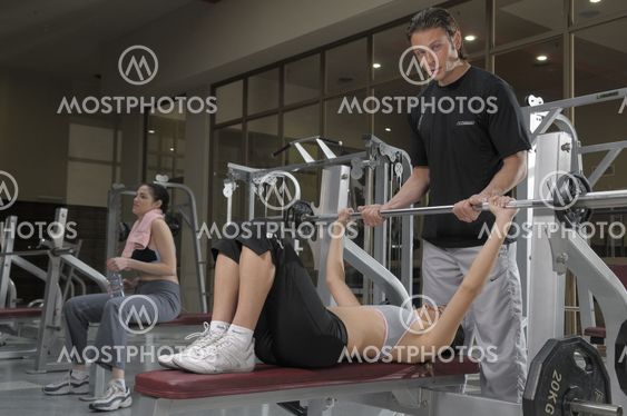 Personal trainer wearing a gray top and black trousers helping to a woman to do weights at gym