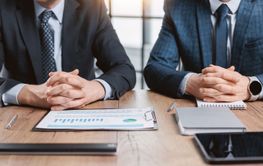 Two men in a suit are sitting at a desk with folded arms...