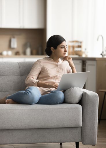 Pensive young female remote worker sit on couch with...