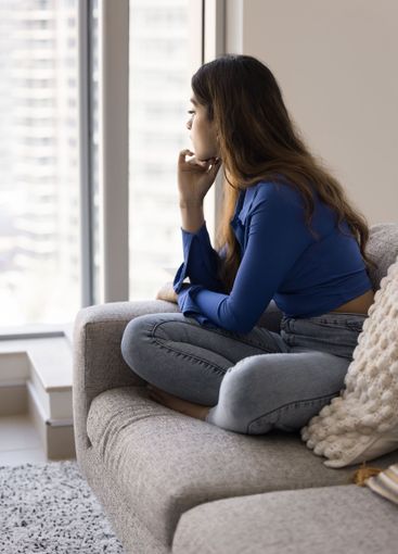 Indian woman sitting on couch, gazing pensively out of...