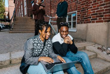 Young female student using laptop while sitting with male...