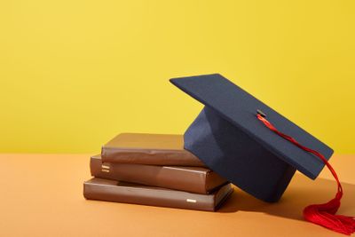 Brown books and academic cap with red tassel on orange...