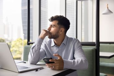 Pensive man holds smartphone sit in coworking looking...