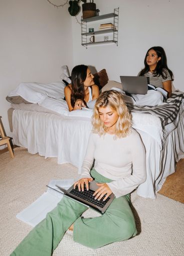 Young woman using laptop while female roommates on bed in...