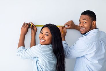 Happy black couple measuring wall with tape-measure,...