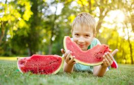 Cute child lying on the grass and eating juicy slice of...