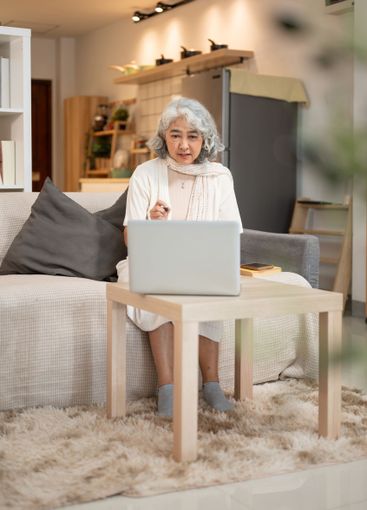 A happy Asian senior woman uses a laptop while sitting...