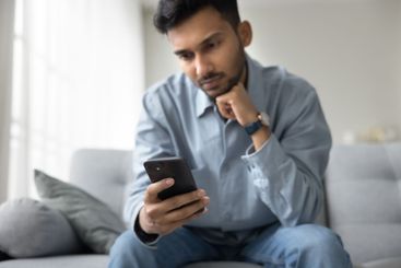 Serious millennial male sit on sofa read news on smartphone
