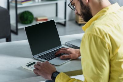 bearded man looking at calculator near laptop with blank...
