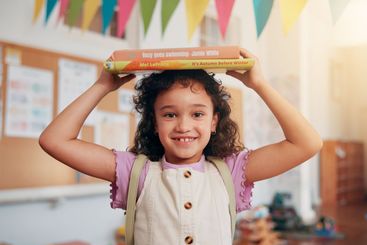 Girl, portrait and books on head in classroom, smile and...