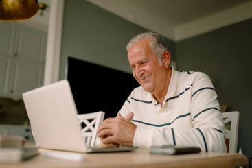 Smiling retired senior man using laptop at dining table...