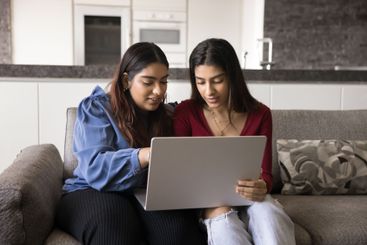 Two Indian women using laptop seated on sofa at home
