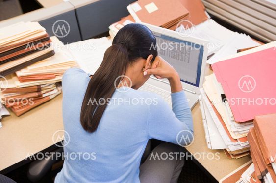Businesswoman in cubicle with laptop and stacks of files