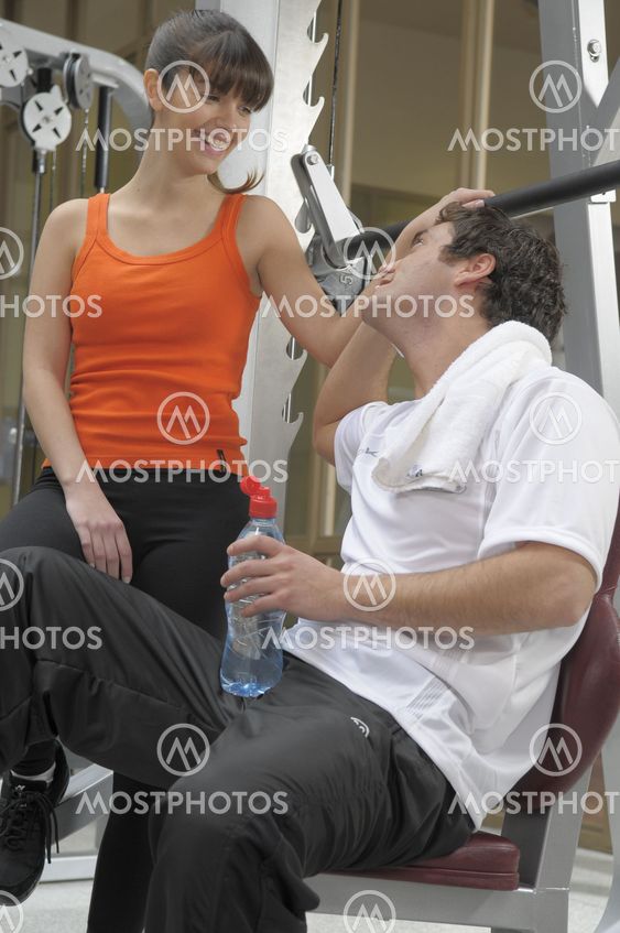 Man wearing a white t-shirt holding a bottle of water sited down on an exercise machine talking to a woman at gym