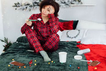Young upset African woman in pajamas is sitting on bed...