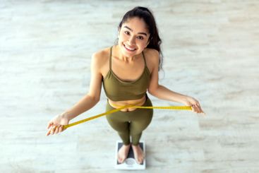 Indian lady measuring waist while standing on scales,...