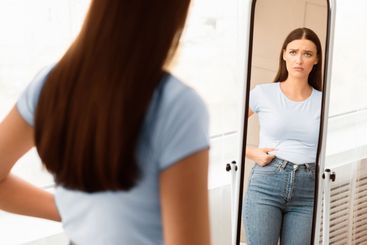 Upset Woman Touching Fat Belly Looking In Mirror Indoor,...