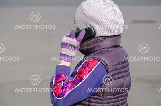 Little girl speaks by mobile phone while standing outdoors.