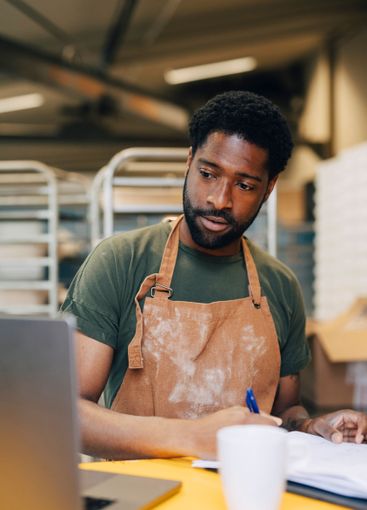 Male baker taking down notes through laptop while working...
