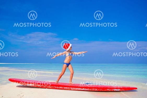 Adorable little girl in Santa hat practice surfing position on surfboard during vacation