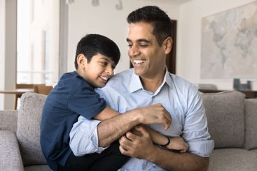 Cheerful Indian dad cuddling teenage son, hugging,...