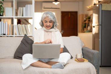 A happy Asian senior woman uses a laptop while sitting...