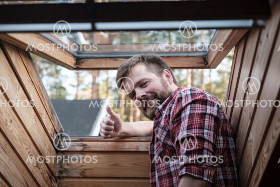 Bearded man stands near an open roof window in attic and...
