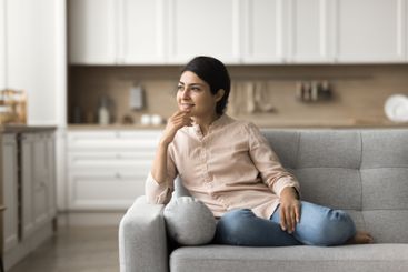 Mindful young lady sitting on couch smiling looking at...