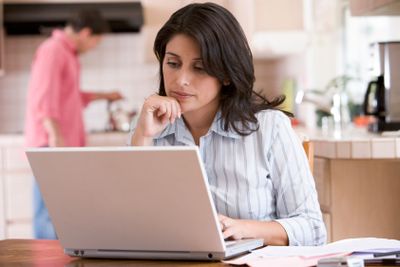 Woman in kitchen with paperwork using laptop with man in...