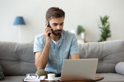 Serious man talk on smartphone using laptop at home