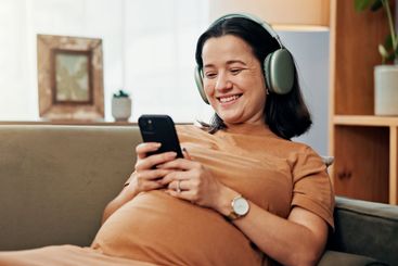 Pregnant woman, phone and headphones on sofa for smile,...