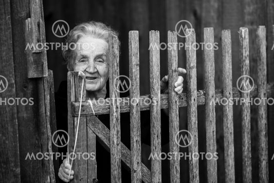 An elderly woman stands behind a wooden fence in the village.