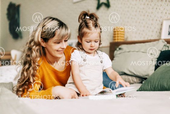 Cute young mom reads a book to her daughter on the bed...