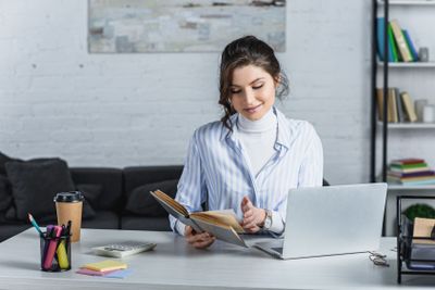 cheerful woman reading book near laptop in modern office