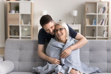 Caring millennial son wrapping shoulders of mom with...