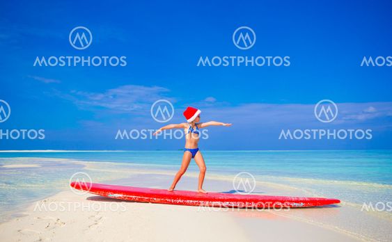 Adorable little girl in Santa hat practice surfing position on surfboard during vacation