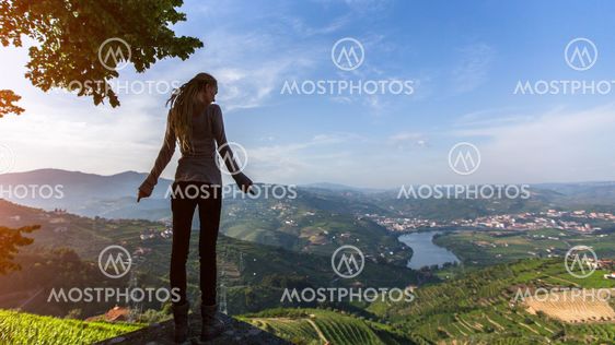 Young woman with blond dreadlocks standing on the edge of a cliff and looks down at on the Douro Valley, Portugal. Looking from the top to Vineyards on a hills.
