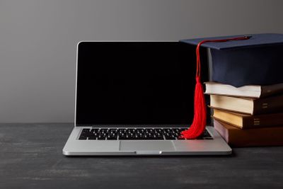 Laptop with blank screen, books and academic cap with red...