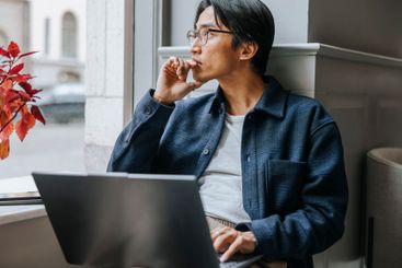 Thoughtful businessman sitting with laptop on window sill...